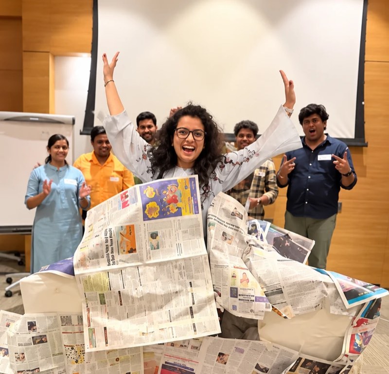 A manager with her arms raised inside a structure her team built from newspaper, the rest of the group applauding behind her